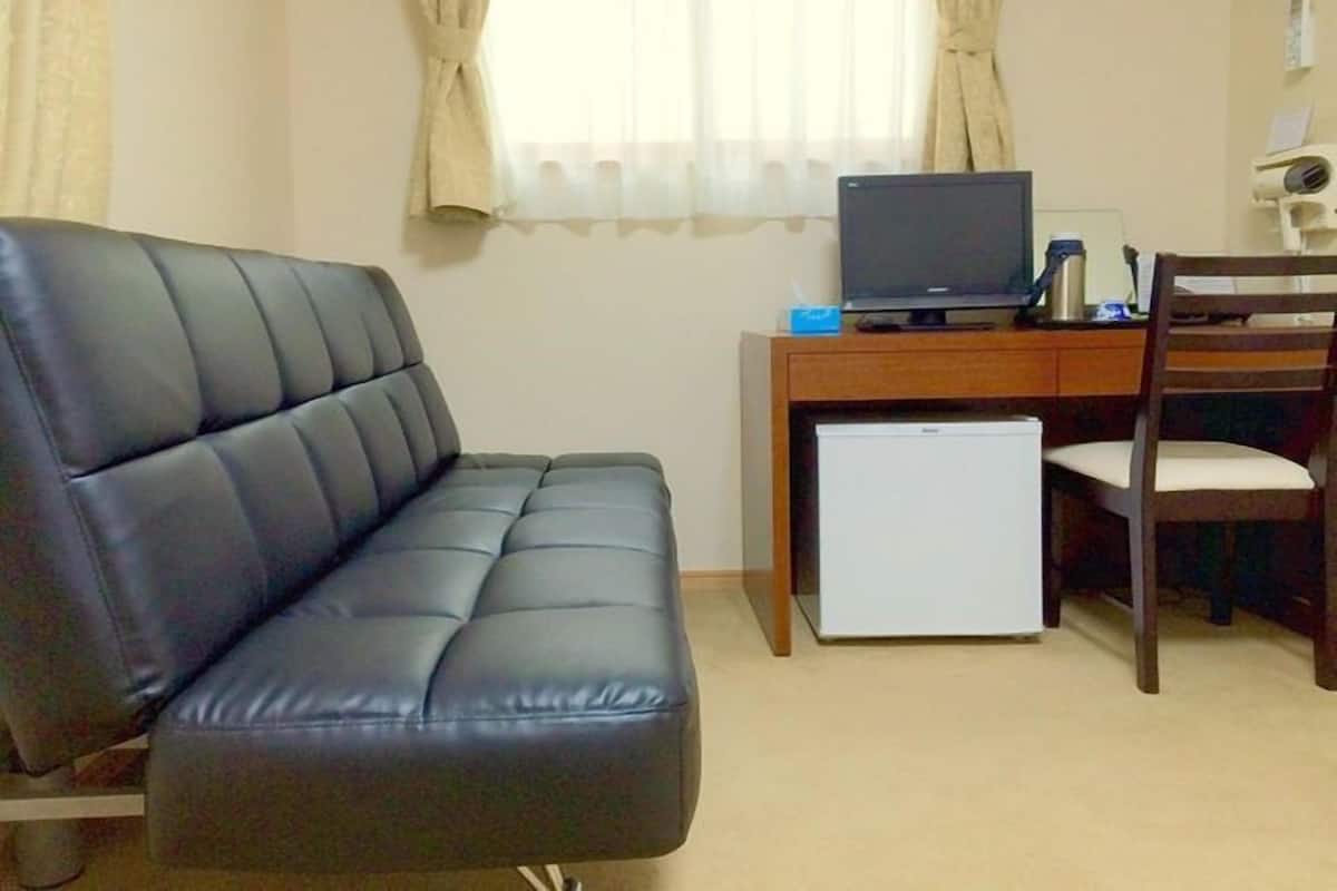 A room with a black leather sofa, a wooden desk with a computer, a small refrigerator, and a chair.