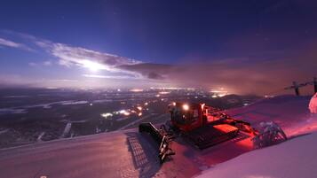 A snowplow on a snowy slope with city lights in the background.