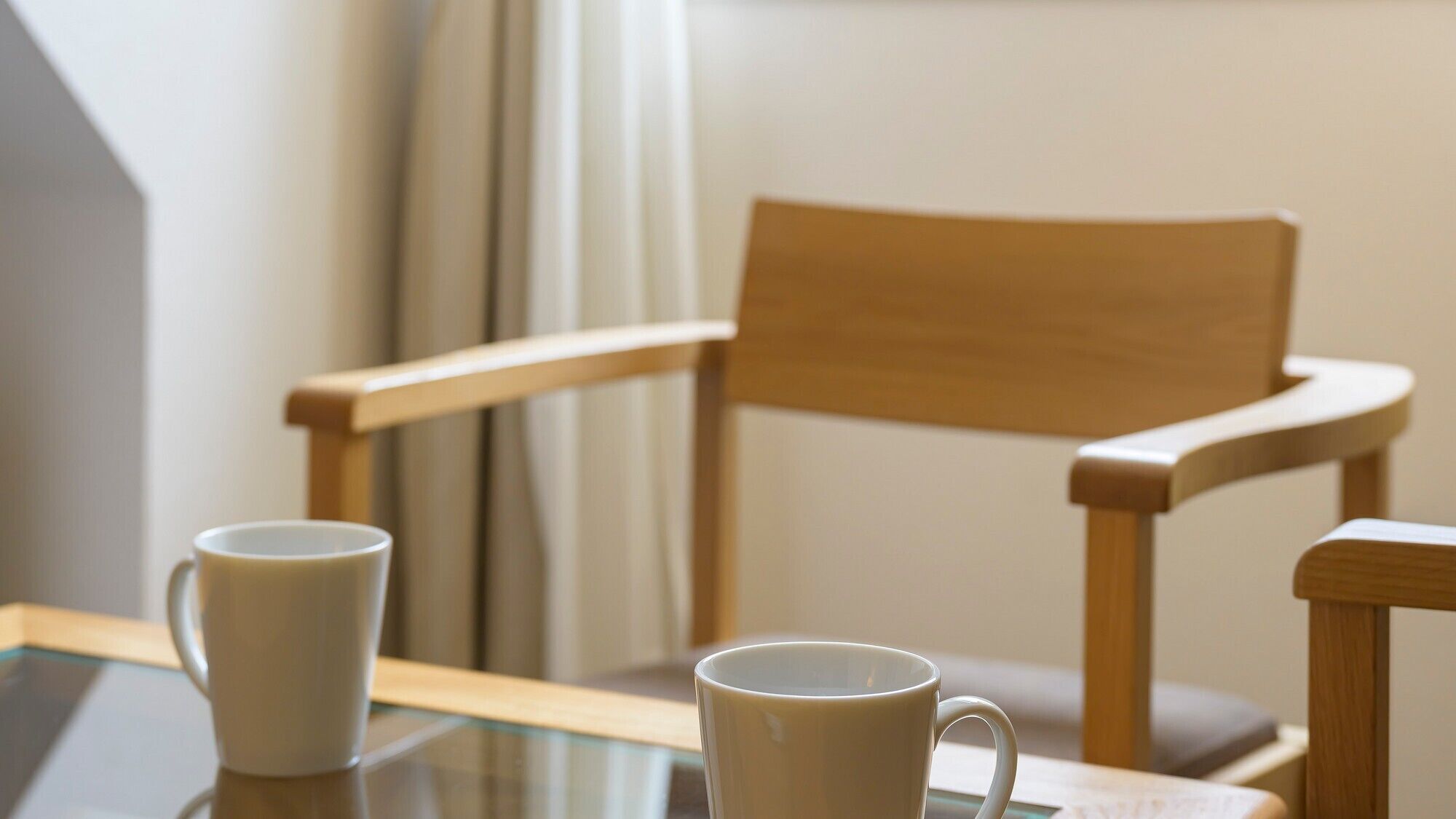 A wooden chair and two white mugs on a glass table.