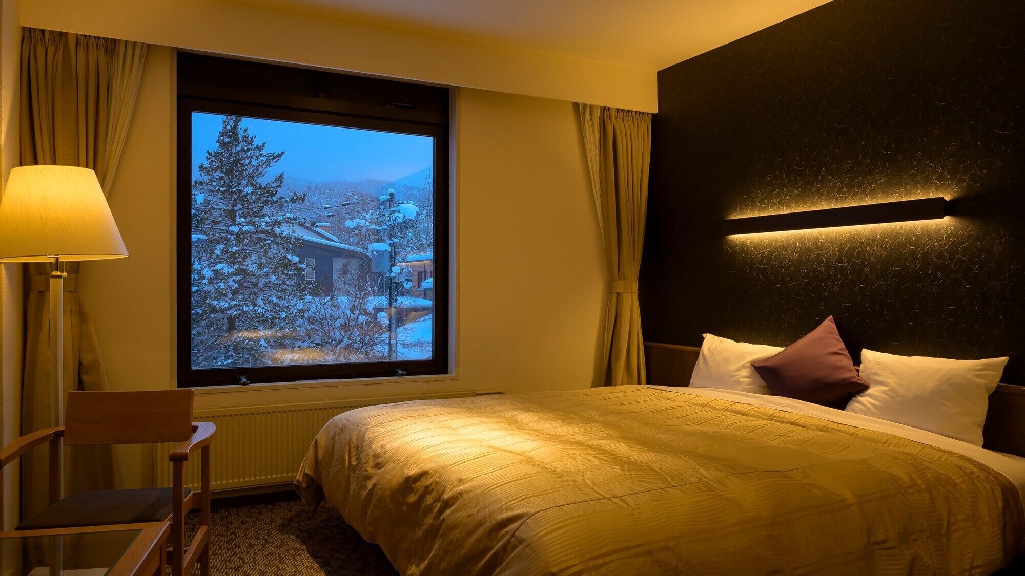 A hotel room with a bed, a chair, a window with a view of a snowy landscape, and a wall-mounted light fixture.