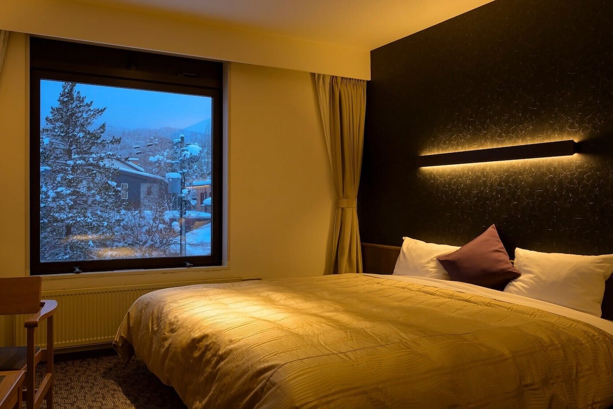 Une chambre d’hôtel avec un lit, une chaise, une fenêtre donnant sur un paysage enneigé et un luminaire fixé au mur.
