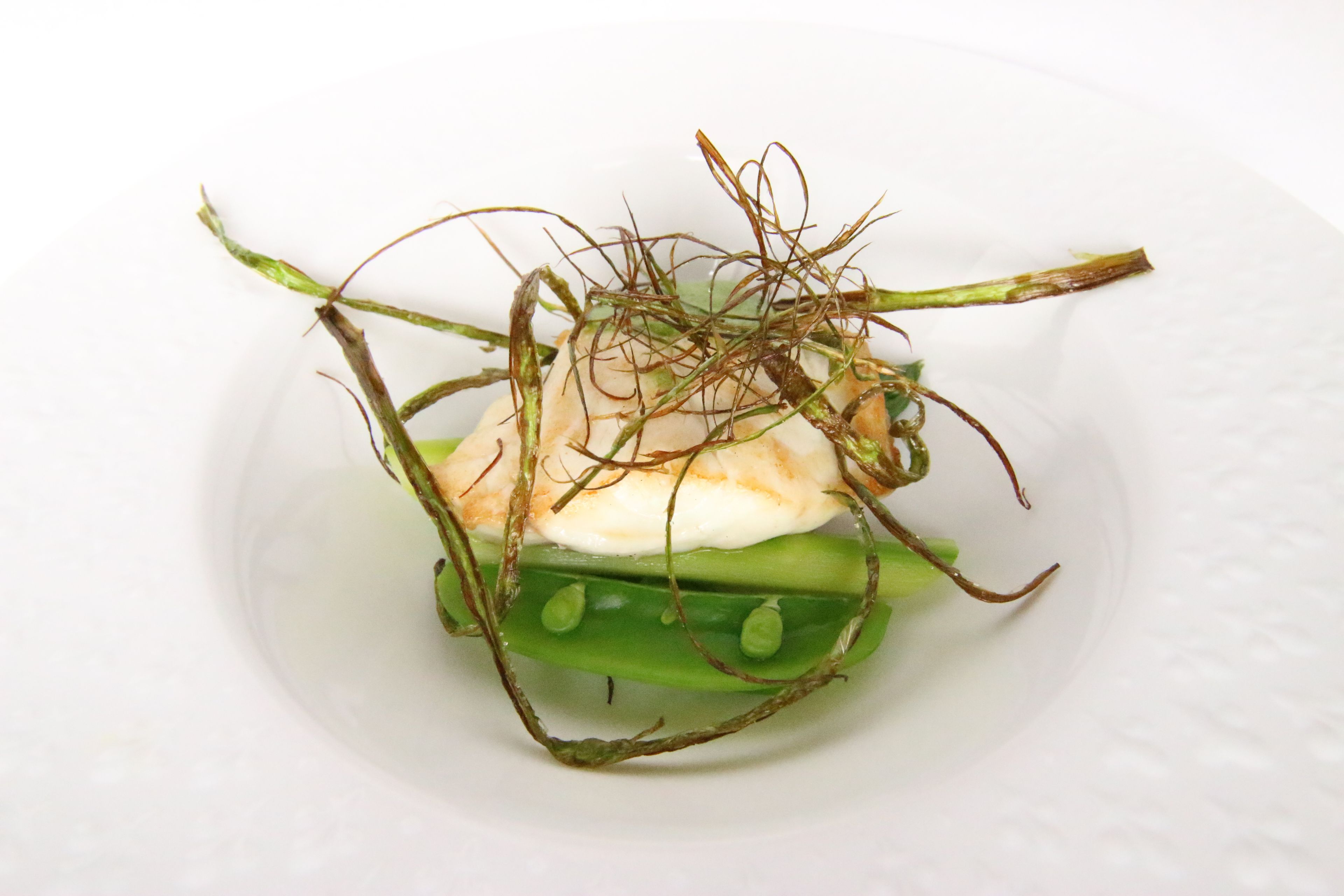 A plate with a white background, containing a green vegetable, possibly a pea pod, and a white creamy substance, likely a dairy product.