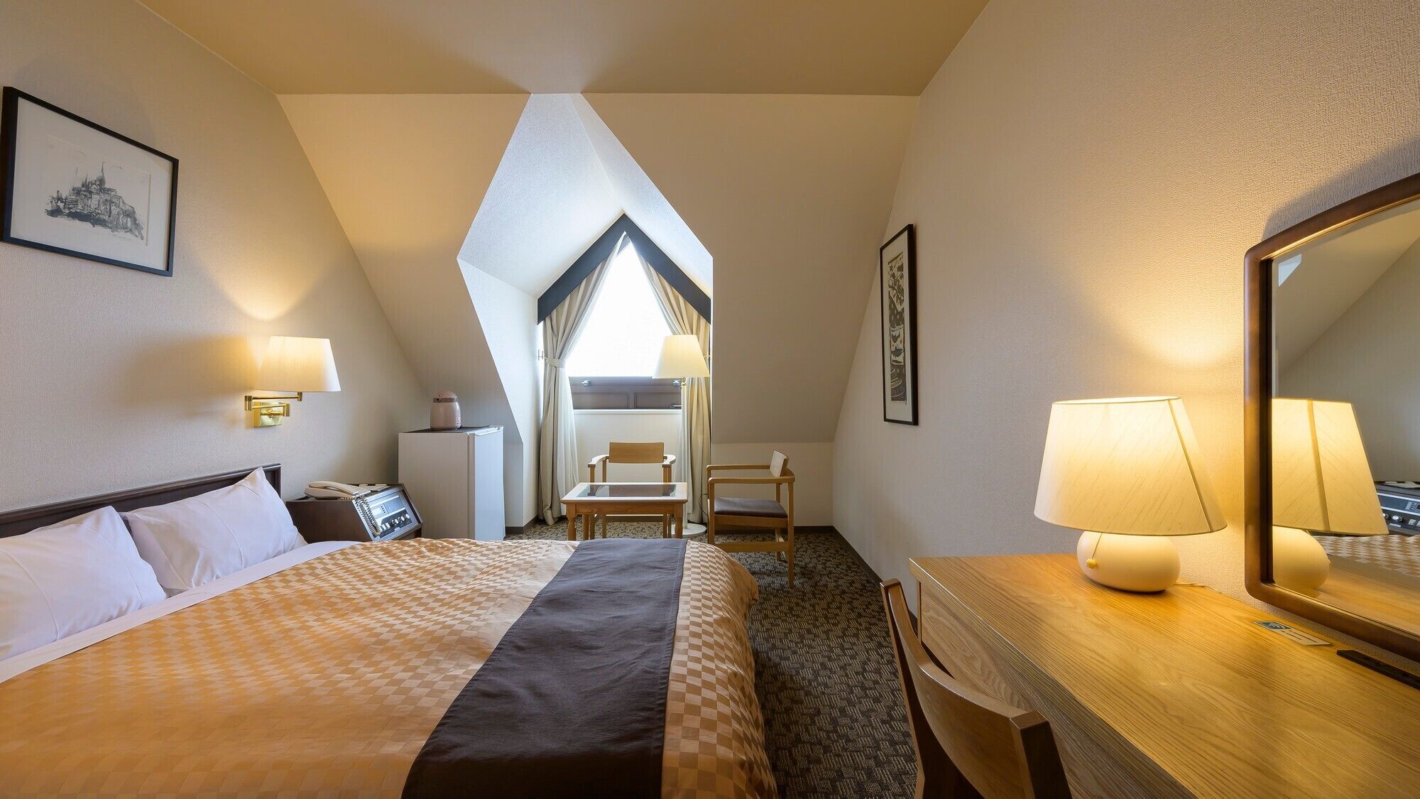 A hotel room with a bed, desk, chair, lamp, and a large mirror.