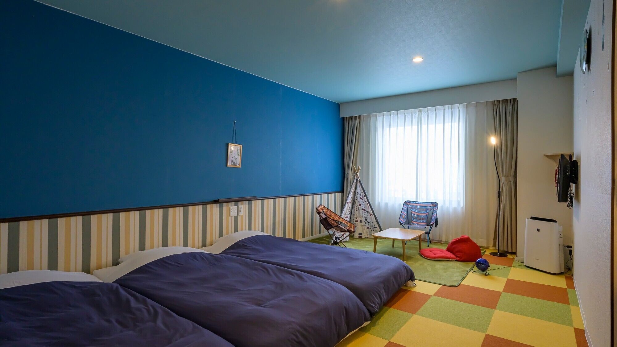 A hotel room with a bed, a chair, a small table, a refrigerator, and a window with curtains.