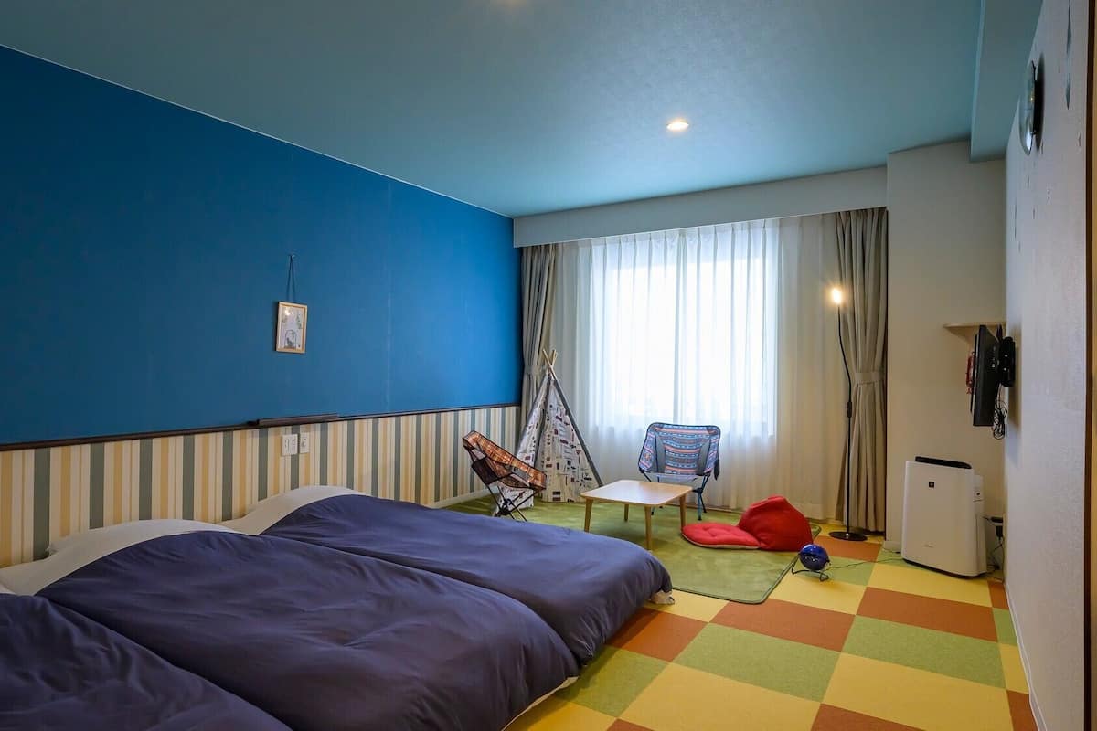 Une chambre d’hôtel avec un lit, une chaise, une petite table, un réfrigérateur et une fenêtre avec des rideaux.