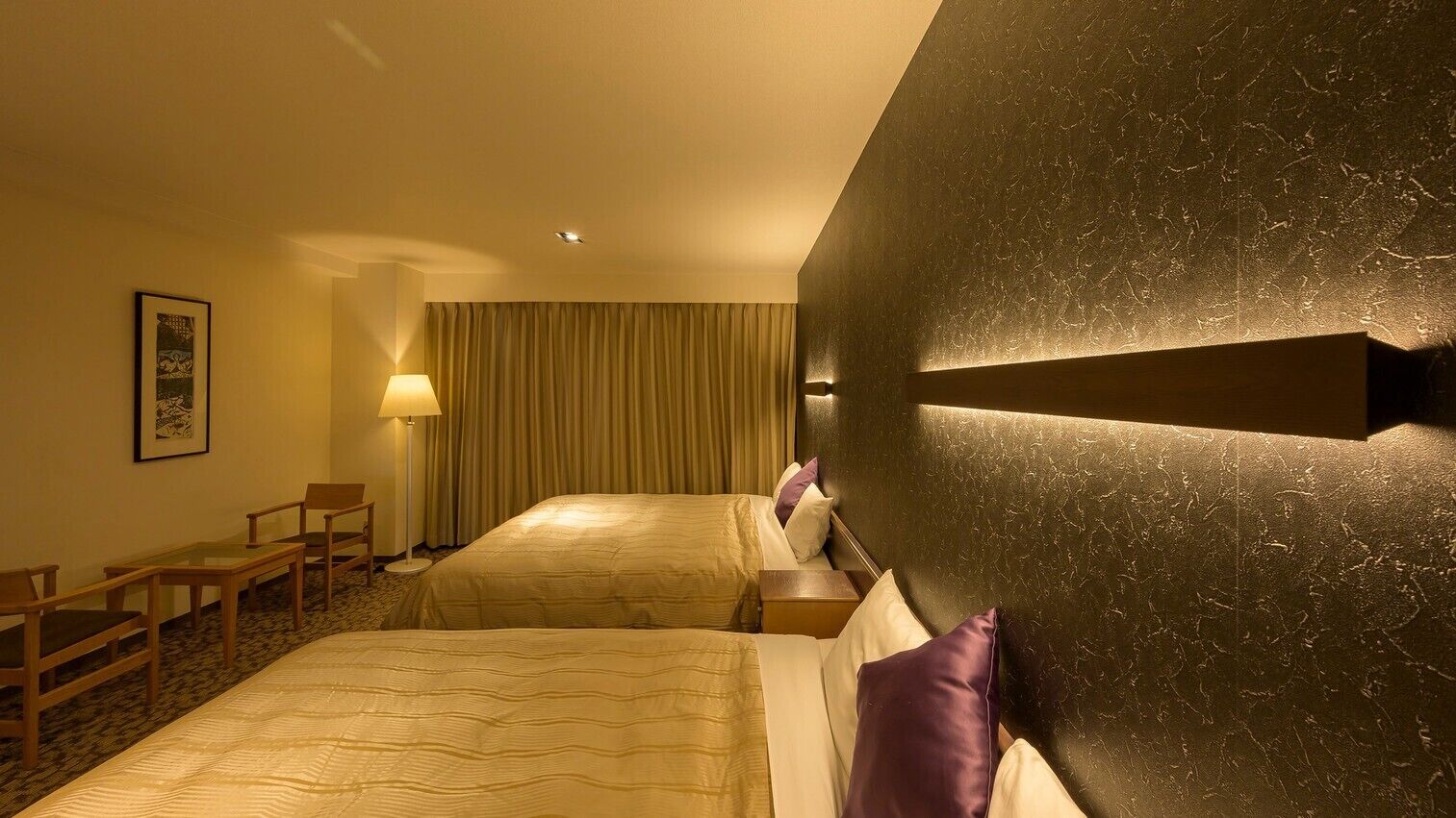 A hotel room with two beds, a desk, a chair, a lamp, and a framed picture on the wall.