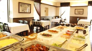 A buffet setup with various food items in metal containers, arranged on a long table in a dining area with chairs and tables.