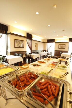 Ein reichhaltiges Buffet mit verschiedenen Speisen in Metallbehältern, präsentiert auf einem langen Tisch in einem Essbereich mit Stühlen und Tischen.