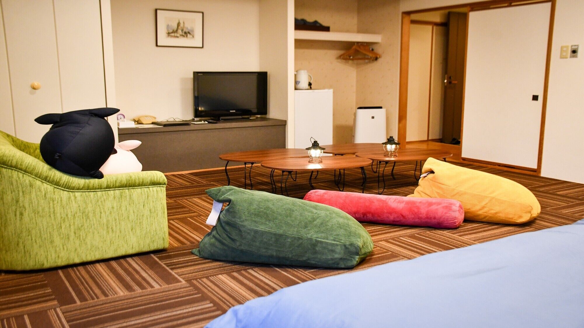 A hotel room with a green sofa, a round dining table with two chairs, a TV, and a small kitchen area.