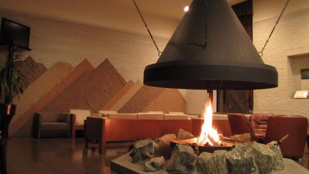A room with a central fireplace, a wooden wall with a geometric pattern, a television mounted on the wall, and a seating area with chairs and a table. - Hotel Edel Warme (Furano)