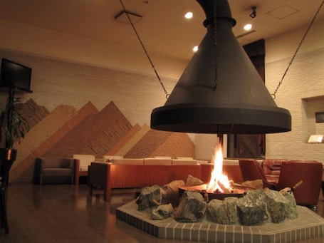 A room with a central fireplace, a wooden wall with a geometric pattern, a television mounted on the wall, and a seating area with chairs and a table.