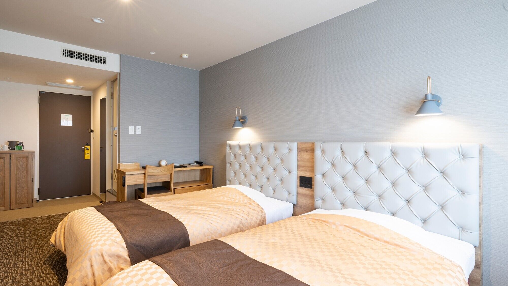 A hotel room with two beds, a desk, a chair, and a lamp.
