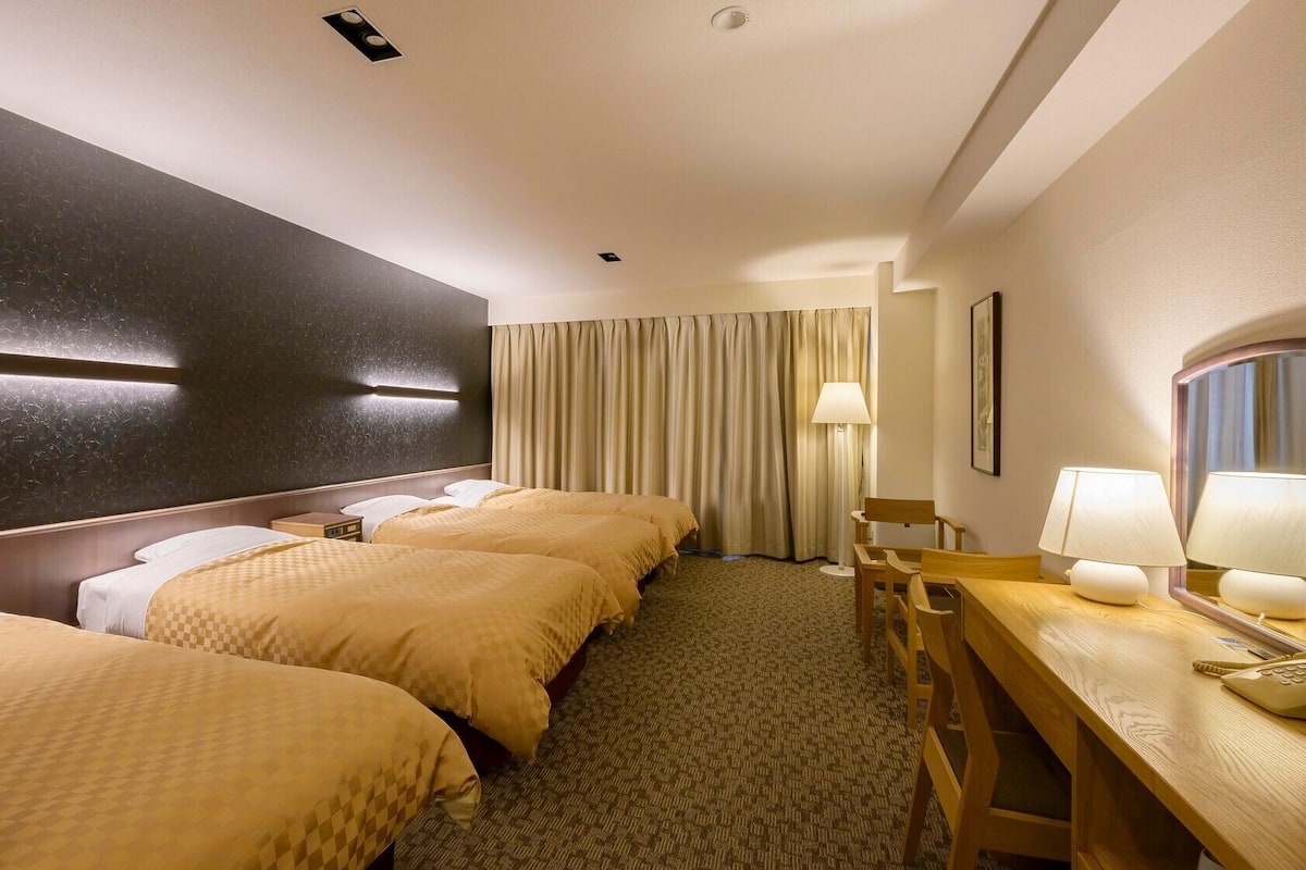 Une chambre d’hôtel avec trois lits, un bureau avec un téléphone, une lampe et un miroir.