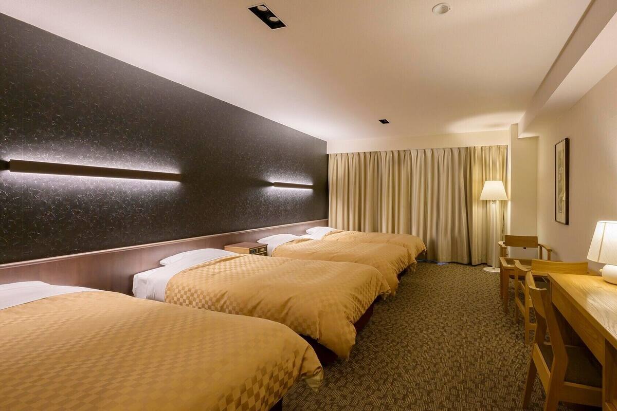 Une chambre d’hôtel avec trois lits, un bureau avec une lampe, une chaise et un grand miroir.