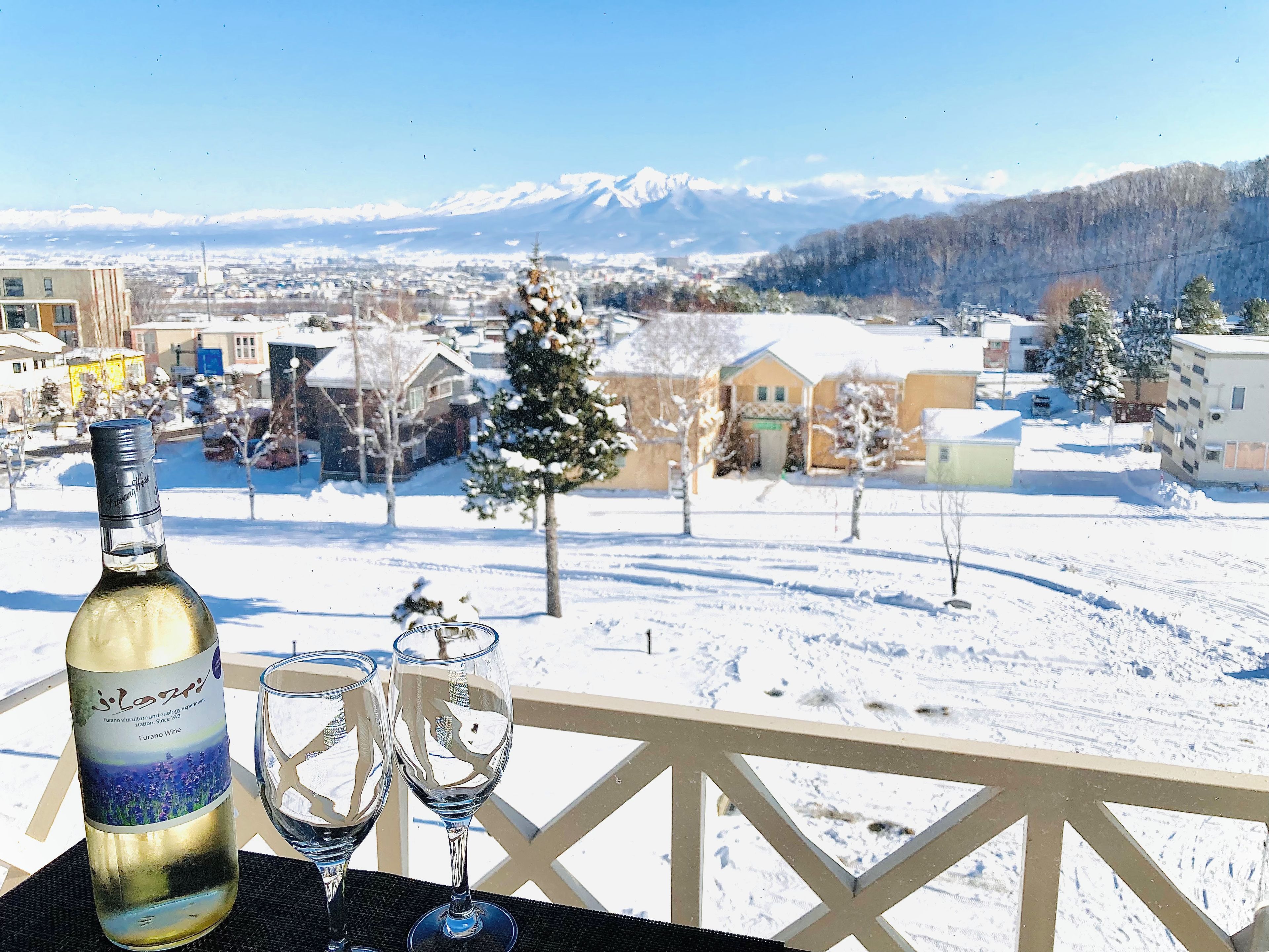 A bottle of wine and two glasses on a balcony with a snowy landscape and mountains in the background.
