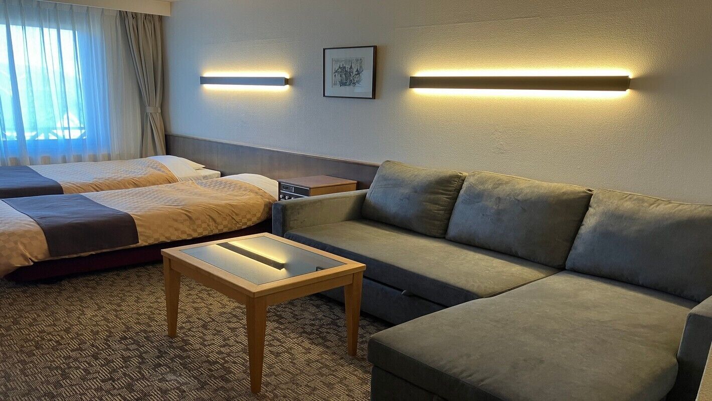 A hotel room with two beds, a sofa, a small table, and wall-mounted lighting.