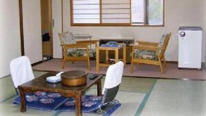 A traditional Japanese-style room with tatami mat flooring, a low table with a bowl, two chairs, a small heater, and a window with shoji screens. - Hotel Masyu (Teshikaga)