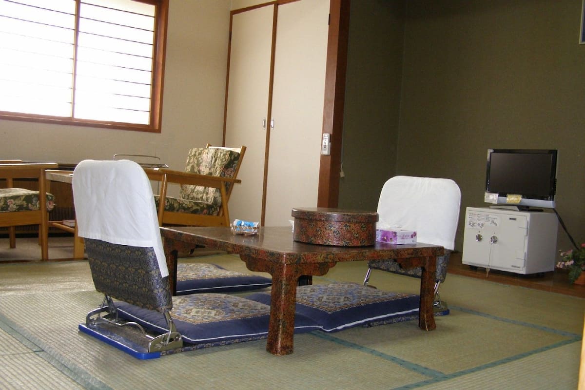 A traditional Japanese room with tatami flooring, a low wooden table, two chairs, a television, and a cabinet.
