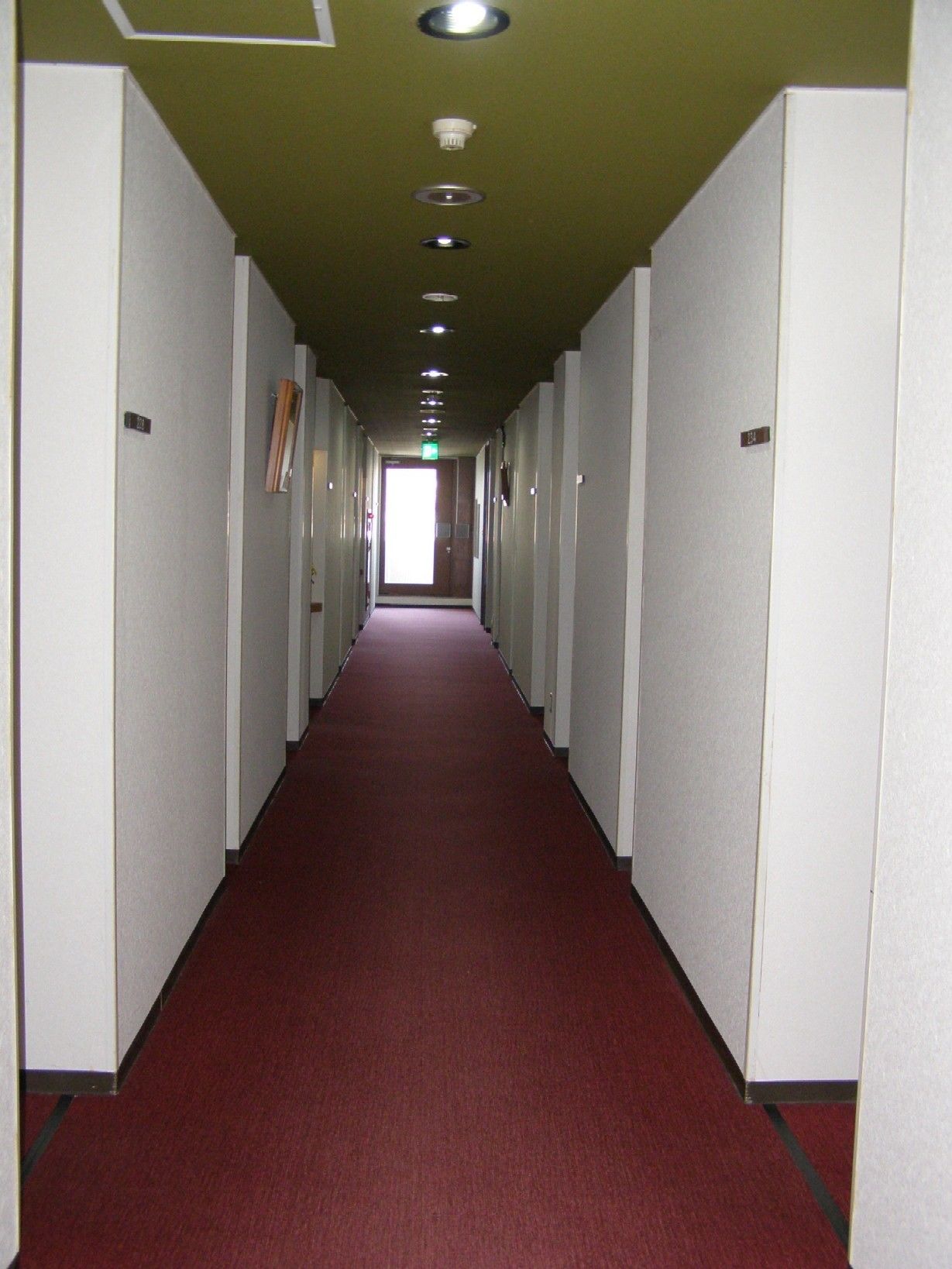 A long, narrow hallway with white walls and a red carpet, leading to a bright exit door.