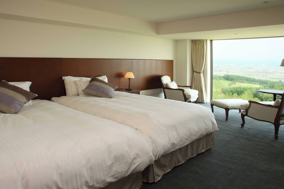 A hotel room with two large beds, a chair, a small table, and a view of the countryside.