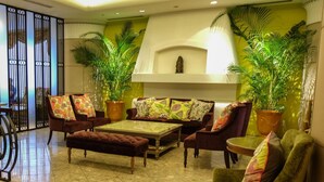 Un vaste espace de vie comprenant un ensemble de canapés, une table basse et deux fauteuils. On y trouve également de grandes plantes en pot et un élément décoratif mural.