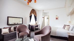 Una habitación de hotel con sofá, dos sillones, una mesa de centro de vidrio, cama, escritorio y candelabro.