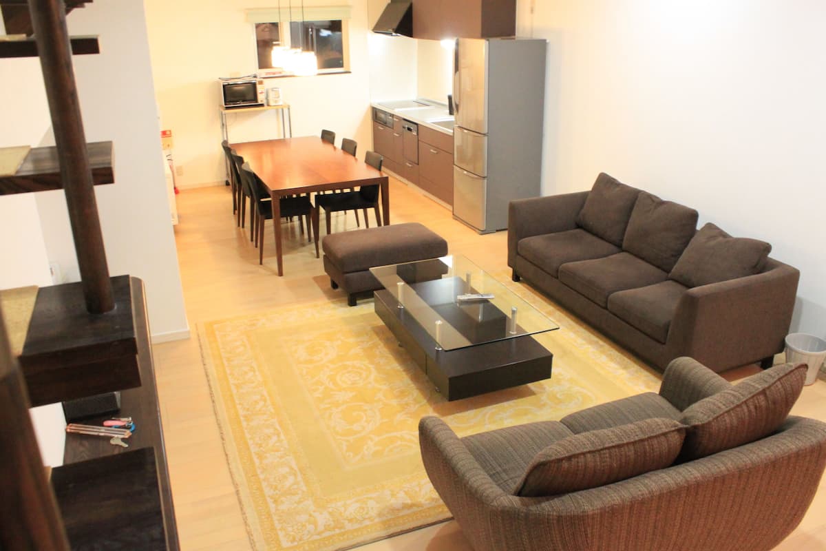 Un salon moderne avec un canapé, un pouf et une table basse. On y trouve également un coin repas avec une table et des chaises, ainsi qu’une cuisine équipée.