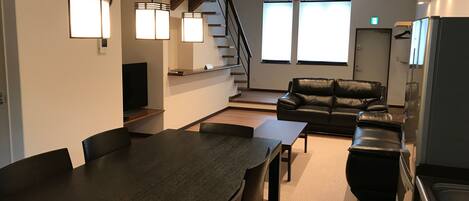 Una sala de estar moderna con un sofá de cuero negro, una mesa de comedor con sillas y una zona de cocina con refrigerador y estufa.