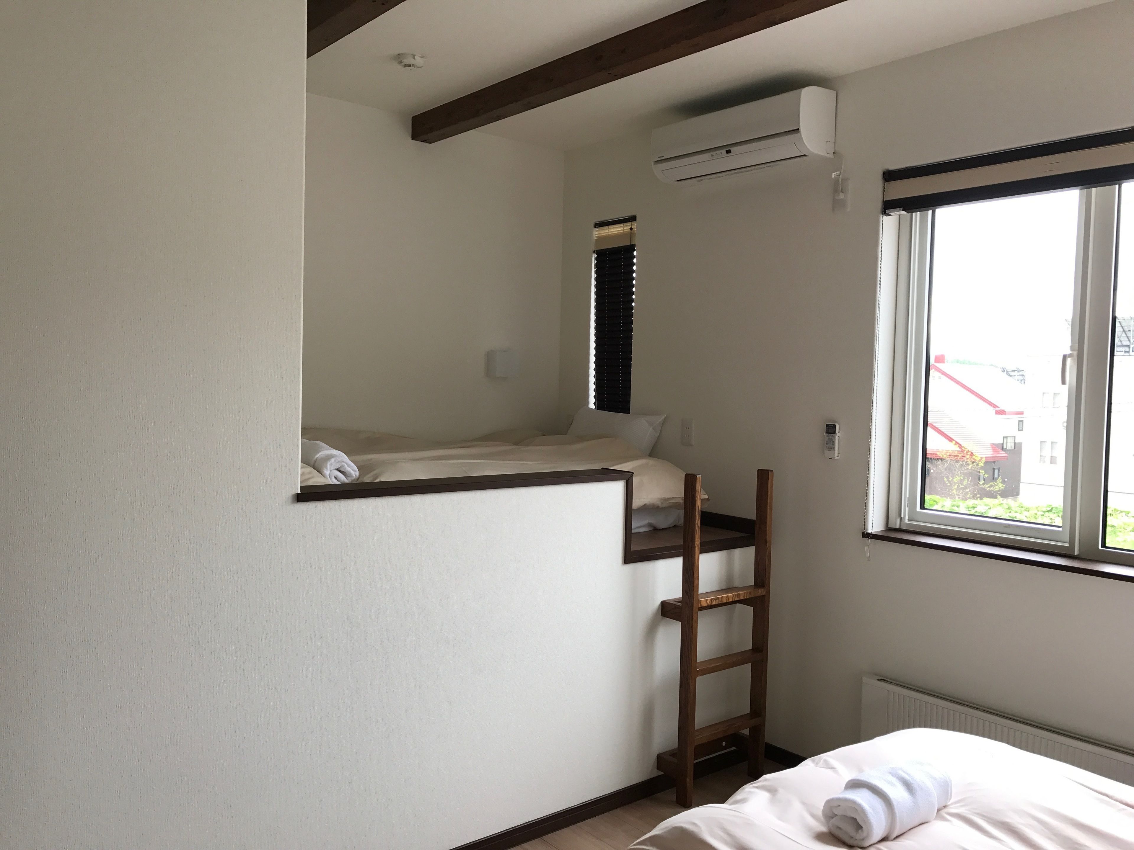 A small, single-bed room with a wooden ladder, a window with blinds, and an air conditioning unit.
