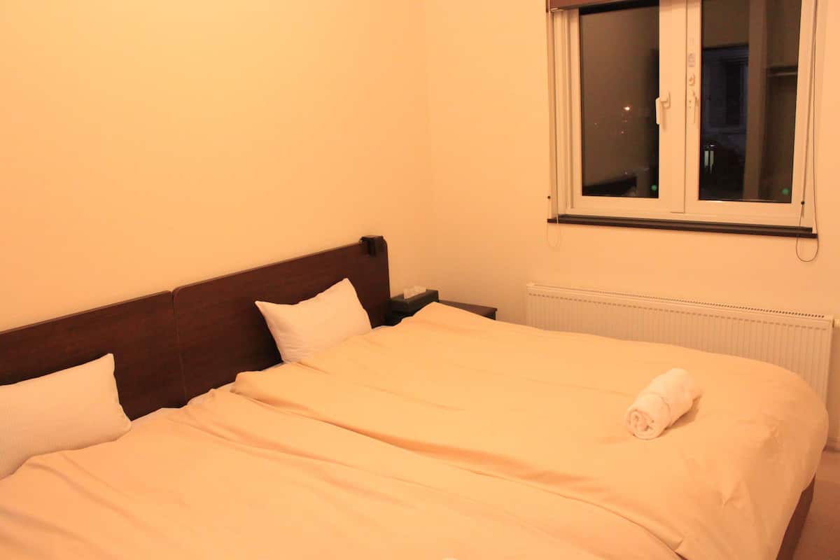 Un lit bien fait, avec du linge de lit blanc et une serviette enroulée, une tête de lit en bois et une fenêtre donnant sur l’extérieur.