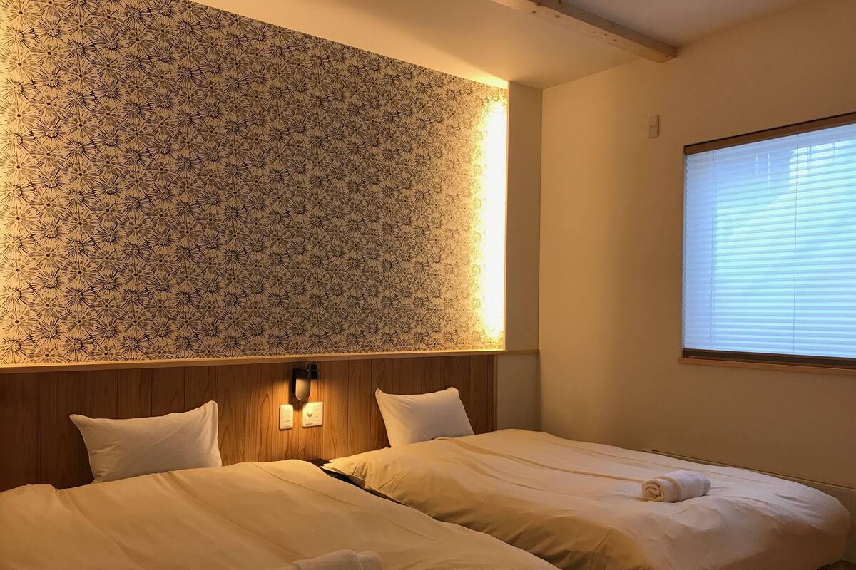 Une chambre d’hôtel avec deux lits, un mur orné d’un motif floral, une tête de lit en bois et une fenêtre avec des stores.