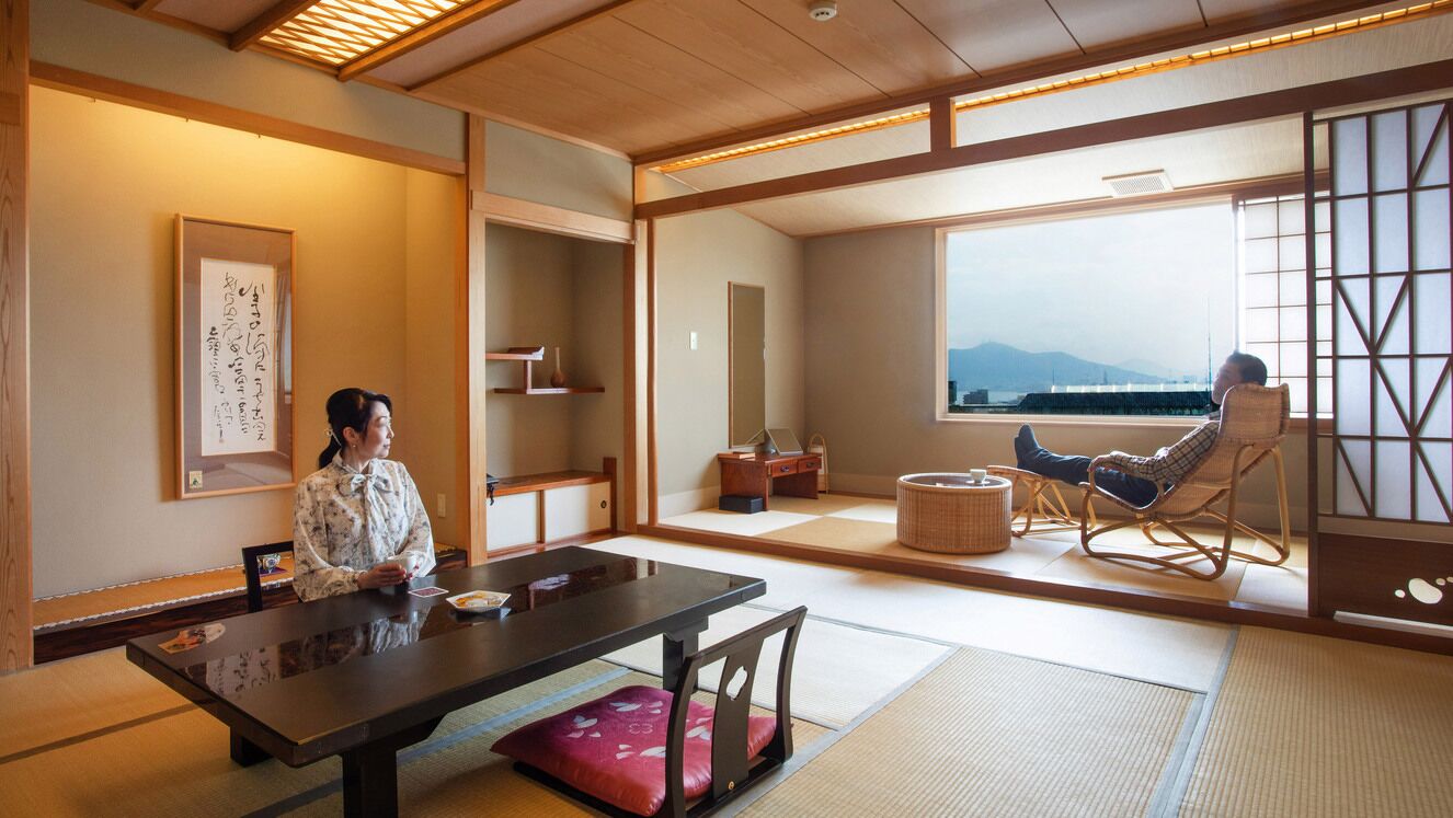 Una habitación japonesa tradicional con suelo de tatami, una mesa baja, una silla acolchada y vistas a las montañas a través de amplias ventanas.