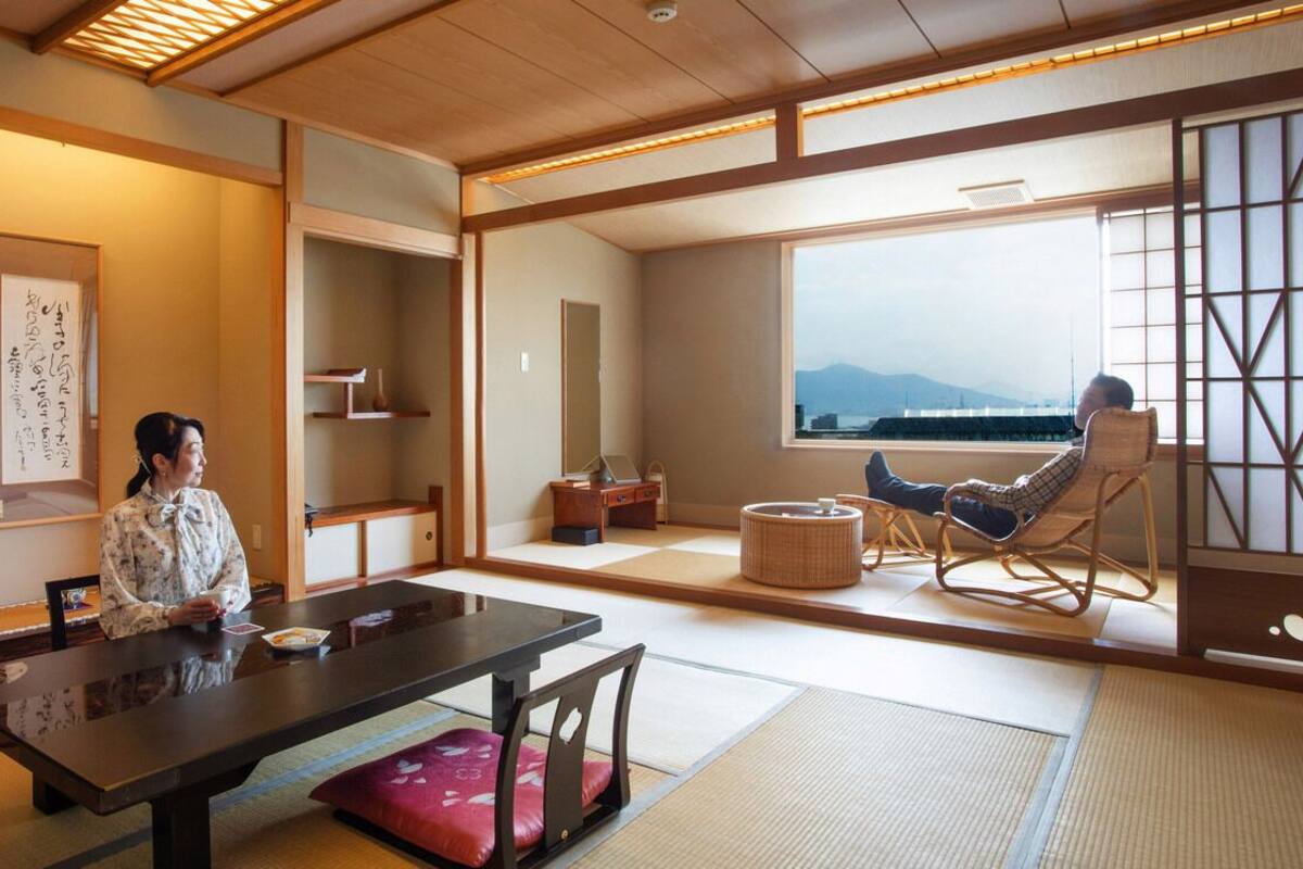 A traditional Japanese room with tatami flooring, a low table, a cushioned chair, and a view of mountains through large windows.