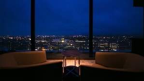 Habitación con un amplio ventanal que da a un paisaje urbano nocturno. Se observan dos sillas y una mesita en primer plano.