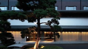 Un edificio moderno con un estilo arquitectónico japonés tradicional, destacando un gran árbol en primer plano y una entrada bien iluminada.