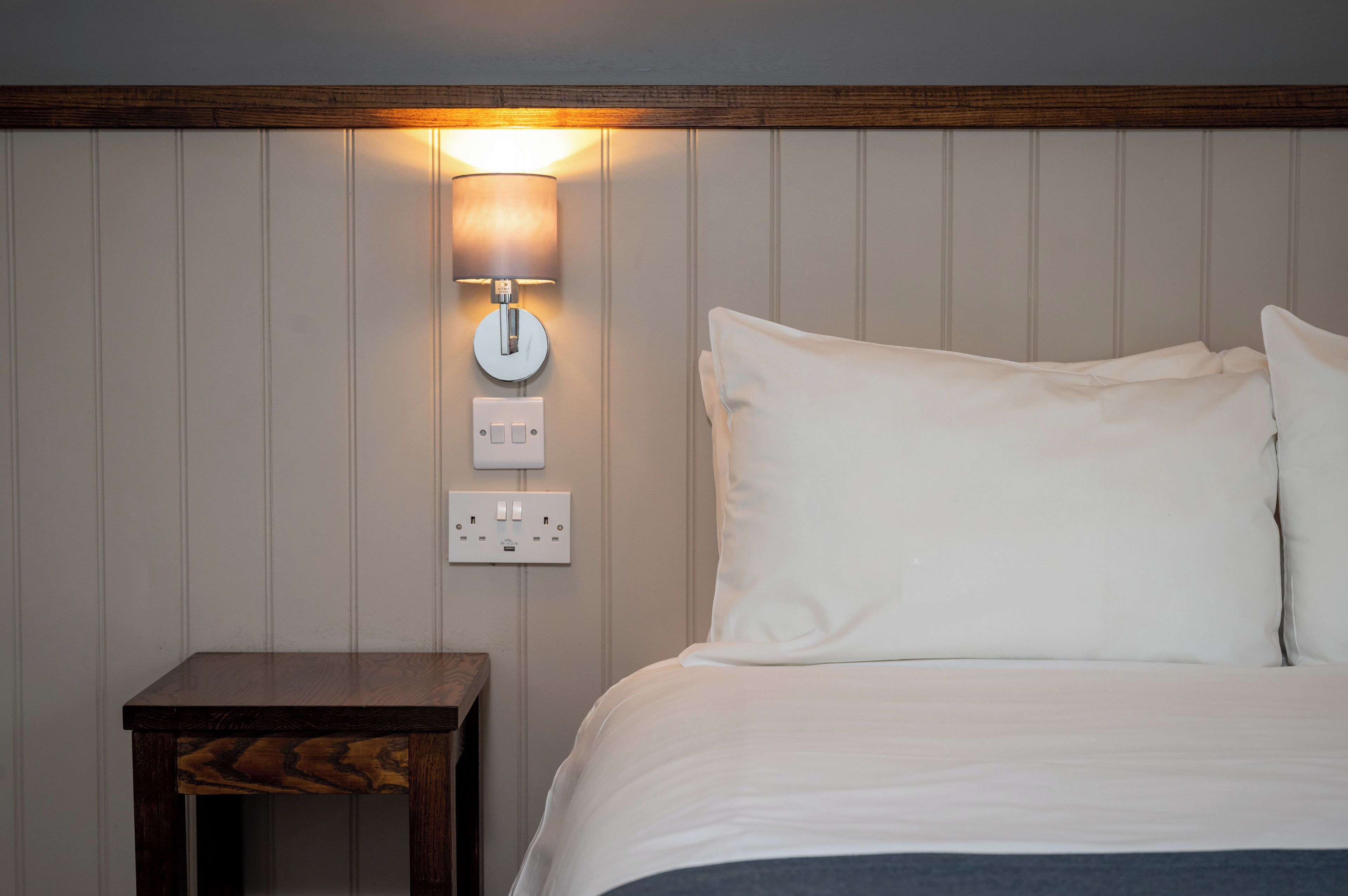 Een keurig opgemaakt bed met witte lakens, een nachtkastje met een lamp, en een wandlamp.