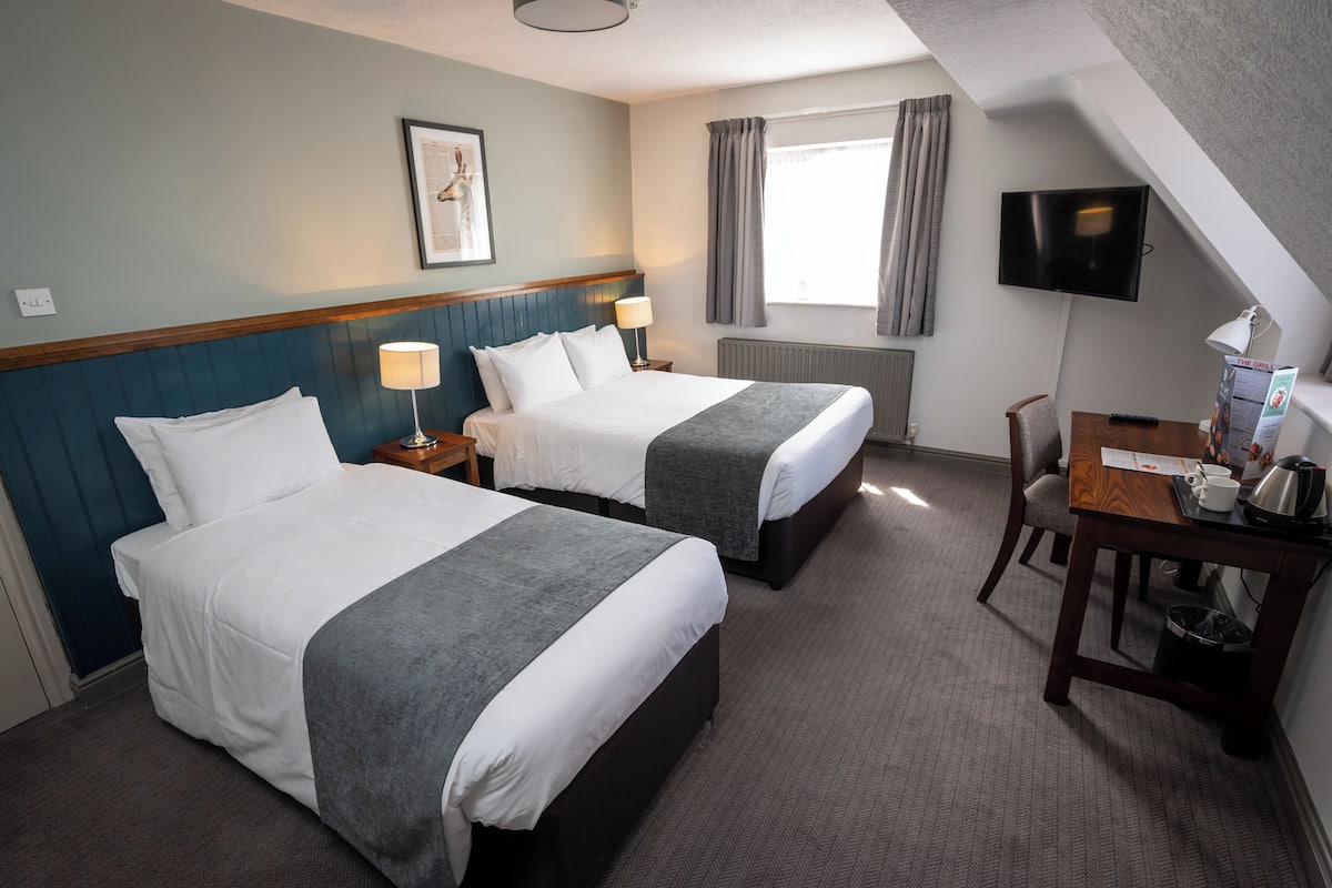 A hotel room with two beds, a desk with a chair, a TV, a window with curtains, and a framed picture on the wall.