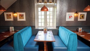 A restaurant with a wooden table and blue cushioned booths, framed pictures on the walls, and a window with natural light. - Cocked Hat Hotel by Greene Kings Inns (Coventry)