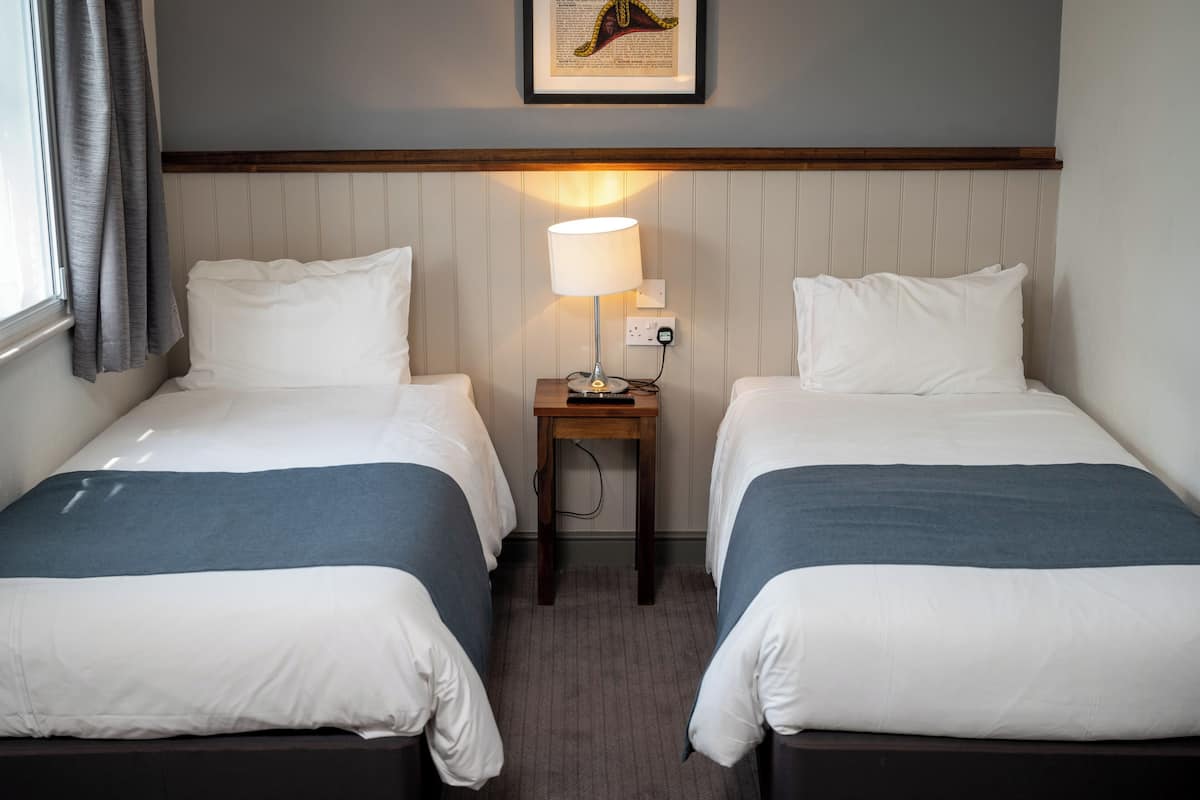 A hotel room with two single beds, a nightstand with a lamp, a framed picture on the wall, and a window with curtains.
