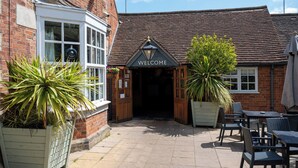 A brick building with a 'WELCOME' sign, outdoor seating, and potted plants.