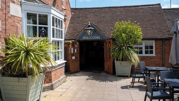 A brick building with a 'WELCOME' sign, outdoor seating, and potted plants. - Cocked Hat Hotel by Greene Kings Inns (Coventry)