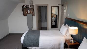 A hotel room with a bed, bedside table with a lamp, a mirror, a door leading to a bathroom, and a shelf with a fan and other items.