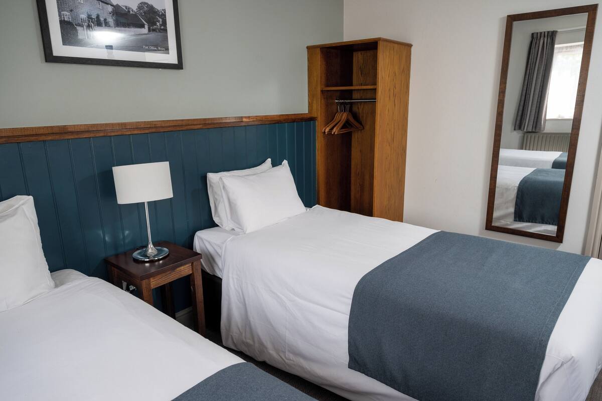 A hotel room with two beds, a wooden wardrobe, a bedside table with a lamp, and a framed picture on the wall.