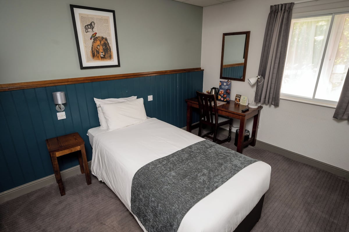 A neatly arranged hotel room with a bed, a desk, a chair, a small table, a lamp, and a window with curtains.