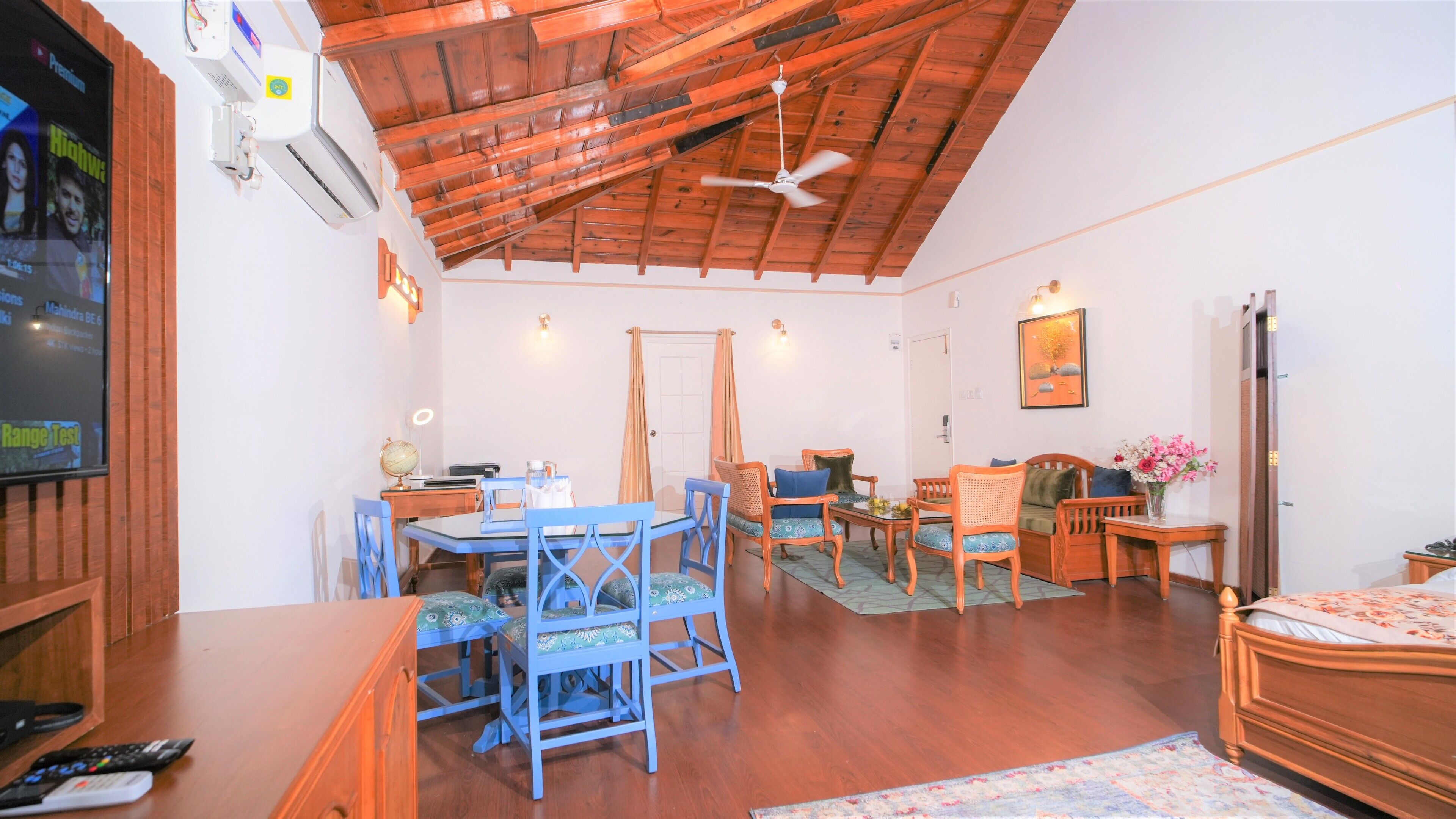 A room with a wooden ceiling, a dining area with blue chairs, a bed, and a TV on the wall.