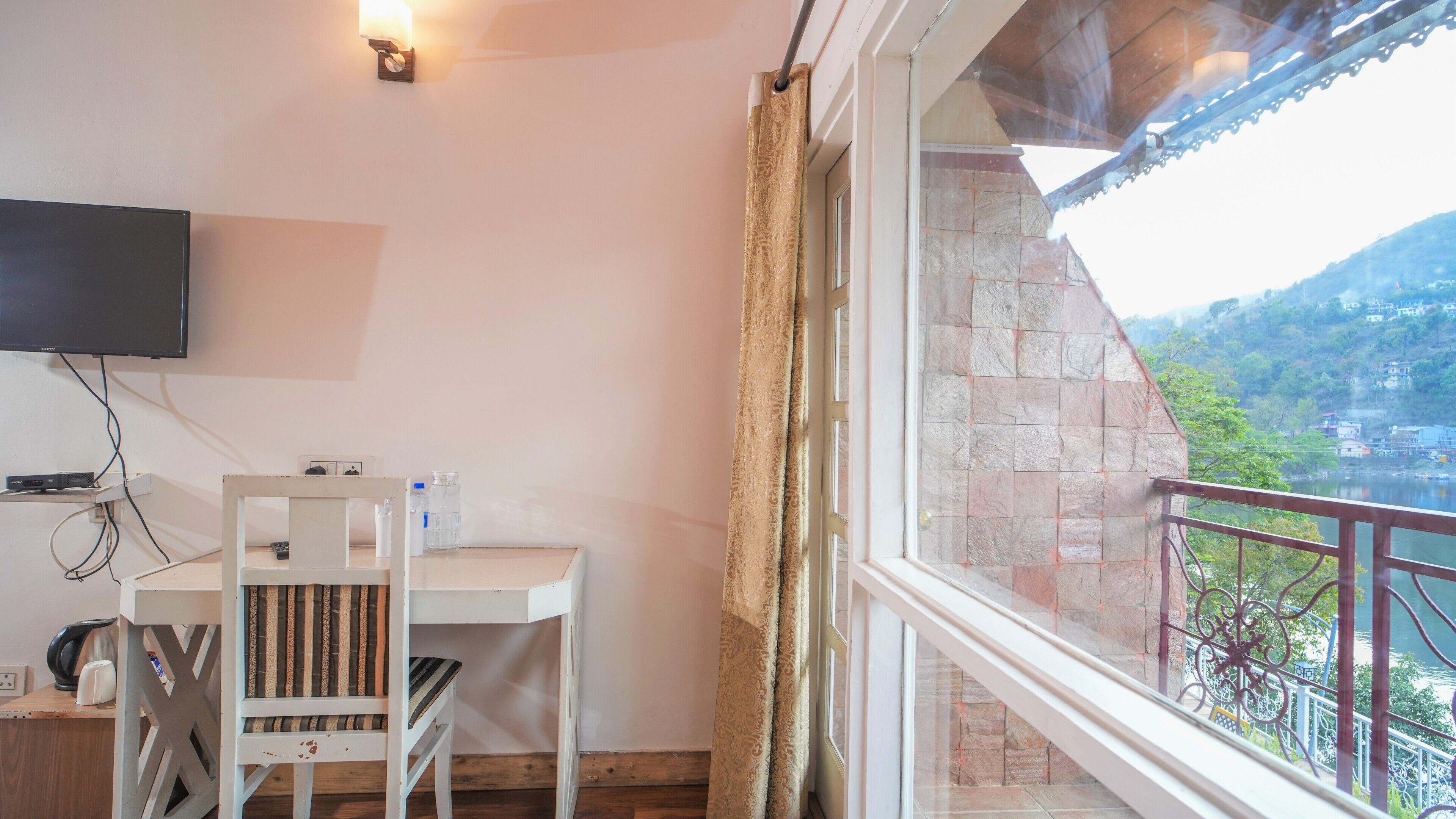 Una habitación con televisión montada en la pared, una mesa con sillas, una ventana con cortinas y vistas a un paisaje montañoso.