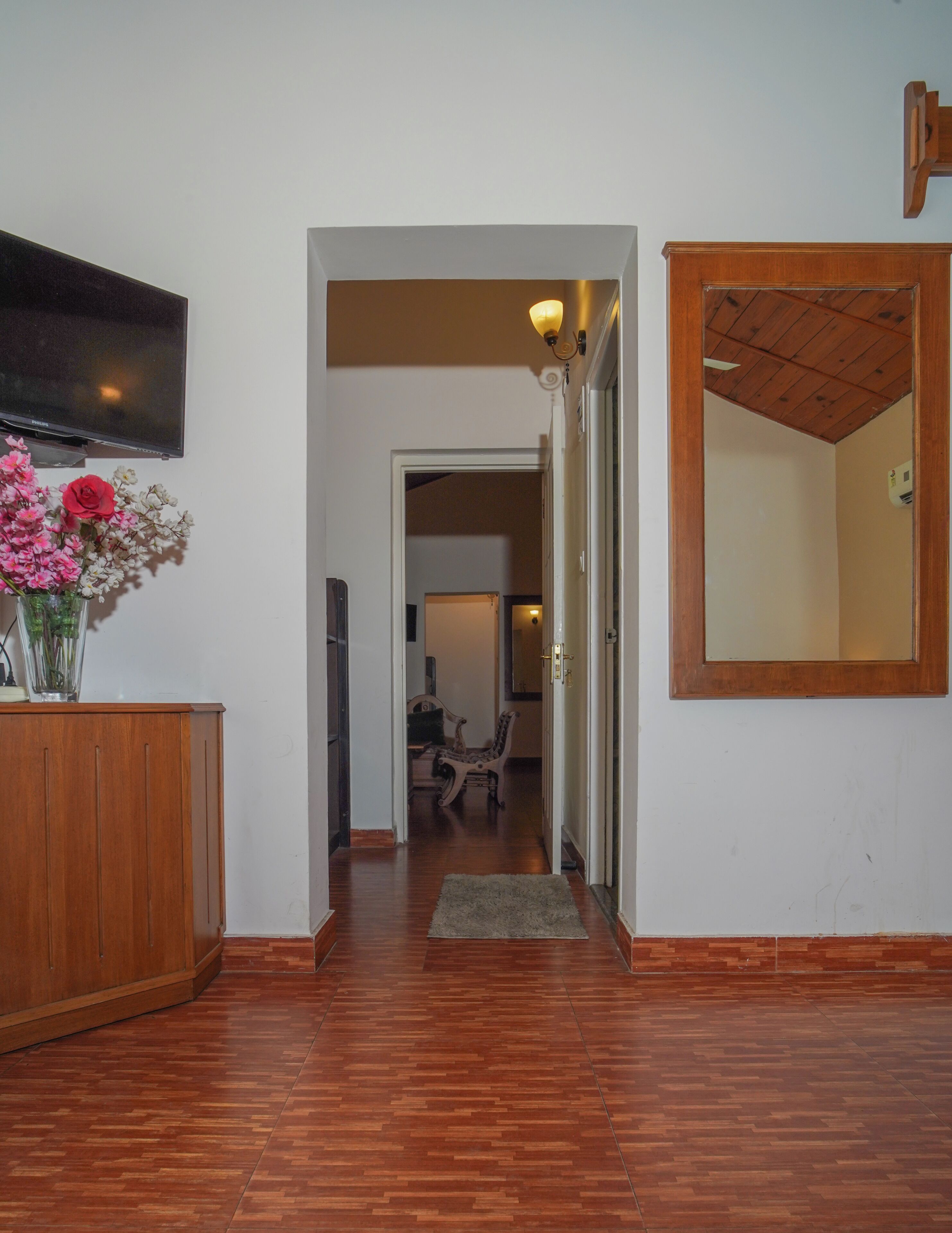 A hallway with wooden flooring, a wooden cabinet, a vase with flowers, a mirror, and a television.