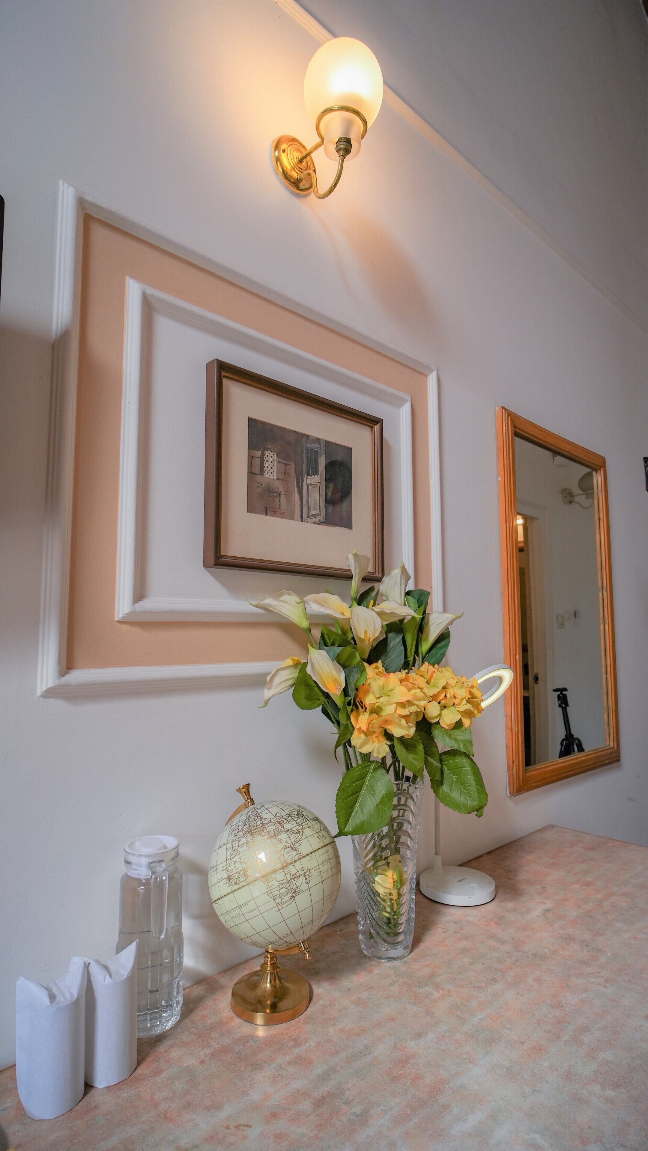 A wall-mounted light fixture, a framed picture, a mirror, a vase with flowers, a globe, and a tissue box.