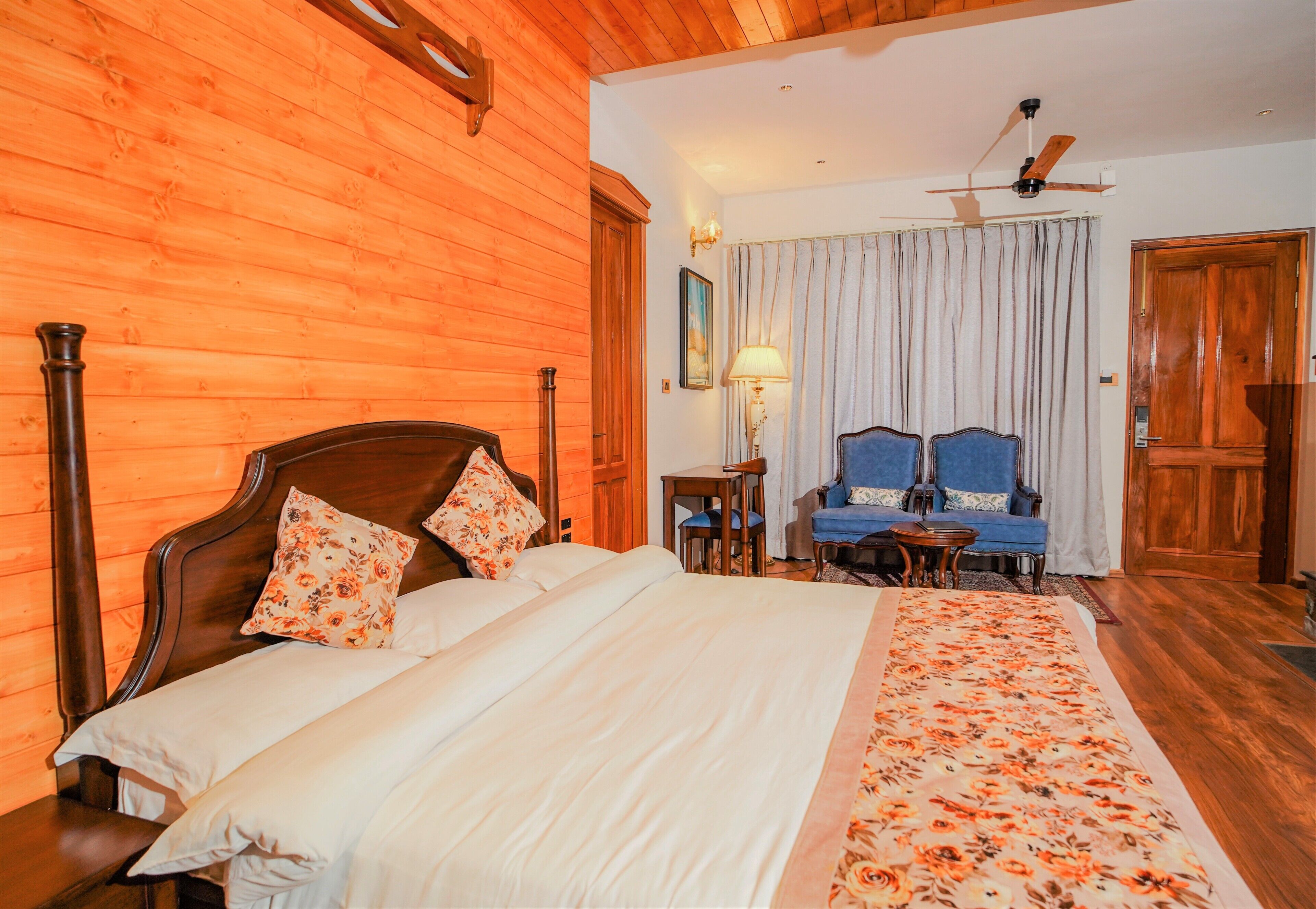A wooden-paneled room with a bed, a chair, a table, and a ceiling fan.