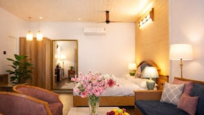 A hotel room with a bed, a sofa, a coffee table with fruit, a vase of flowers, a mirror, and a ceiling fan. - Neelesh Inn - Luxury Lake View Hotel With Swimming Pool On Bhimtal Lake (Nainital)